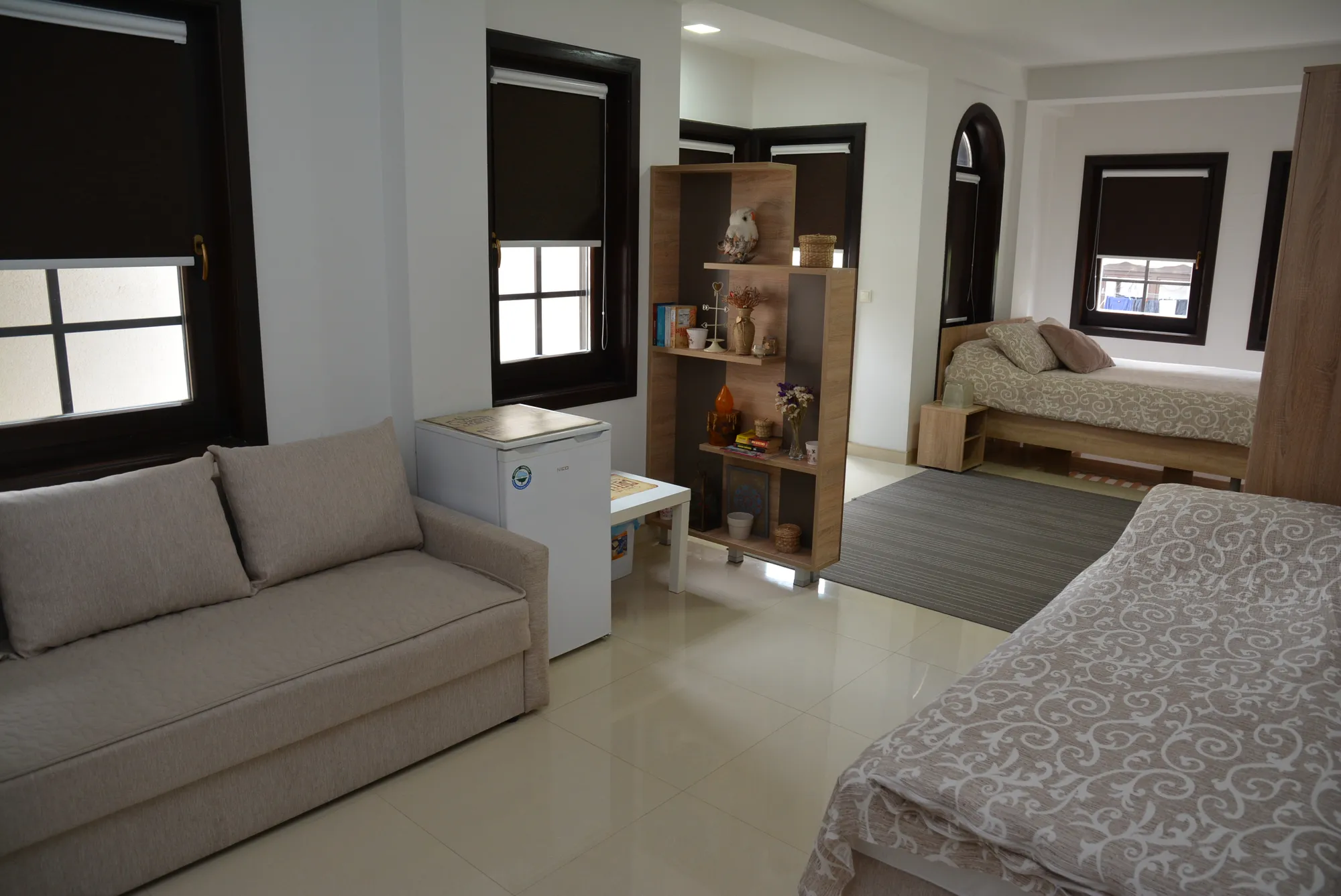 A bright studio room with a sofa, two beds and tall shuttered windows